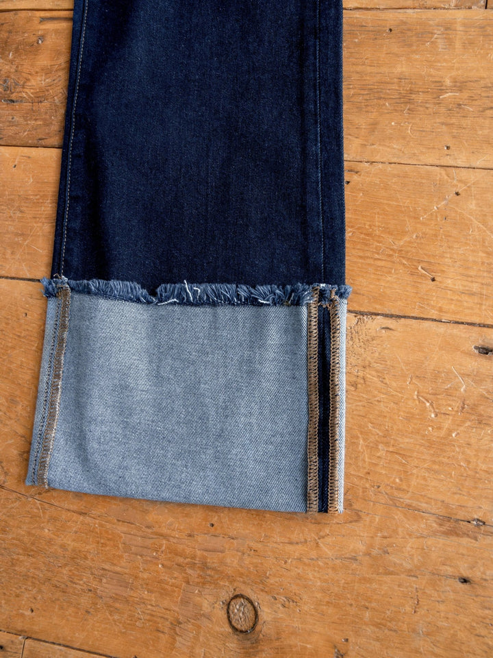 A close-up of The Carson High Rise Wide Leg Jean by Savannah Sevens western life{&}style in dark wash denim, showing a frayed hem folded up to reveal the lighter inside, set on a wooden floor.