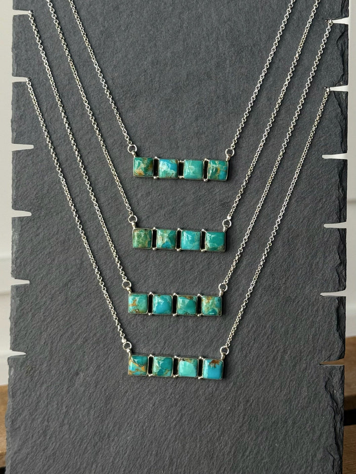 Four Casco Turquoise Bar Necklaces by Savannah Sevens western life{&}style, each featuring rectangular Kingman turquoise stones and arranged in descending order of stone count, are displayed on a textured dark gray stand. These sterling silver necklaces showcase the distinct appeal of the bar necklace design with unique turquoise accents.