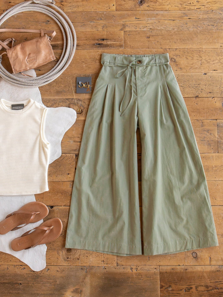 Flat lay of a casual outfit: Savannah Sevens western life{&}style Castana Sage Palazzo Crop Pants, sleeveless cream ribbed top, tan sandals, small tan crossbody bag with round rope strap, gold hoops—all on a wooden floor.