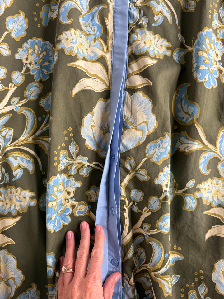A hand touches the Castell Olive Floral Dress by Savannah Sevens western life{&}style, showcasing its blue and cream floral pattern on an olive green background, a central button placket, and light blue lining beneath.