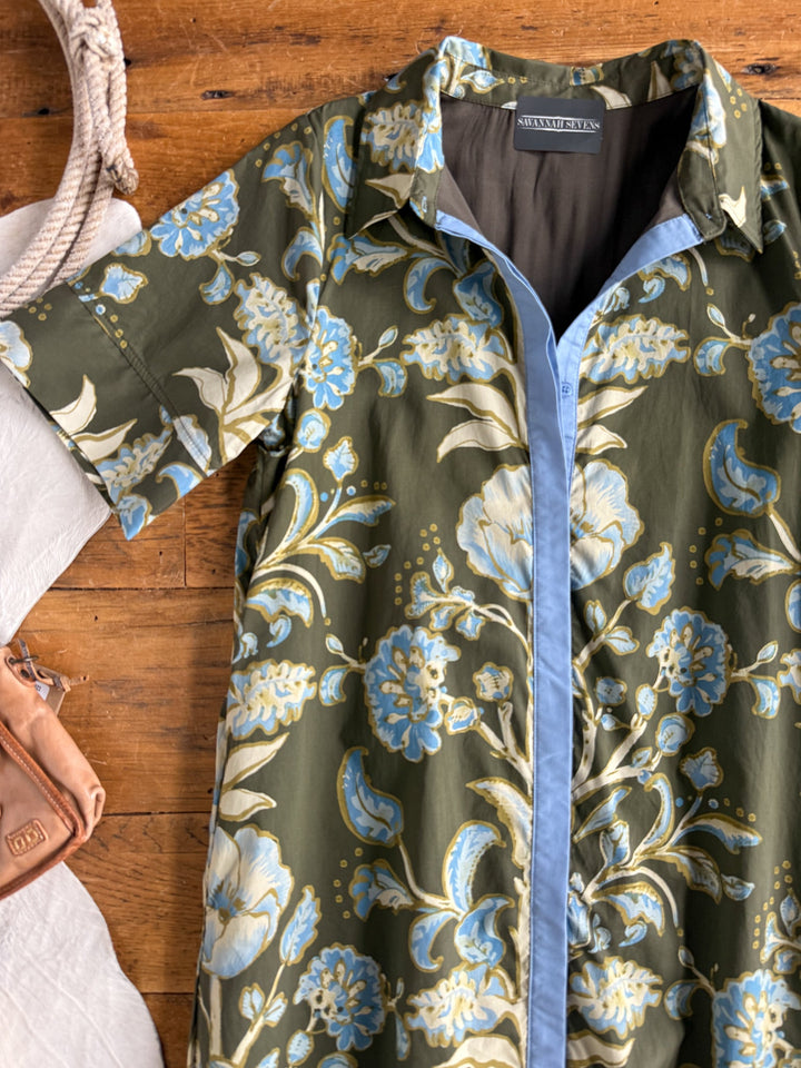 The Castell Olive Floral Dress by Savannah Sevens western life{&}style, featuring blue, white, and yellow floral patterns on an olive green background and short sleeves with a button-up front, is laid on a wooden surface next to a tan purse, white fabric, and a rope.