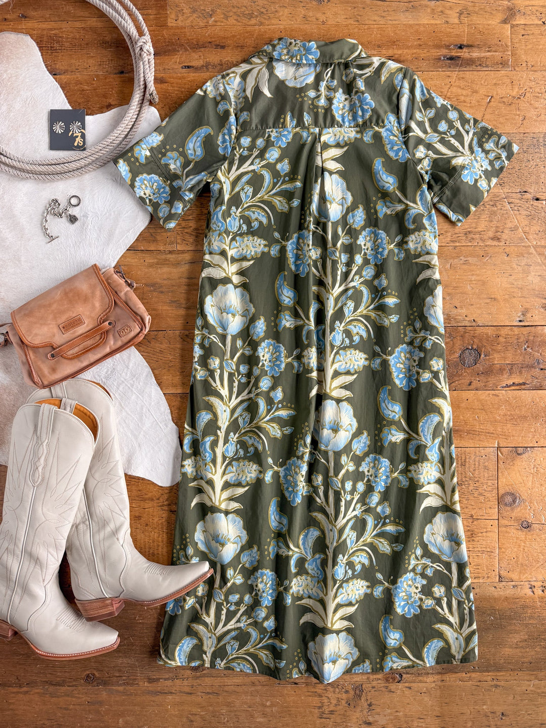 The Castell Olive Floral Dress by Savannah Sevens western life{&}style is laid out with white cowboy boots, a tan purse, silver jewelry, and a coiled rope, all arranged on a rustic wooden floor.