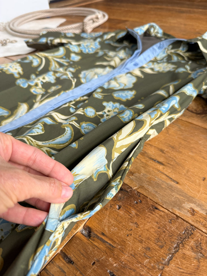A hand holds the hem of the Castell Olive Floral Dress by Savannah Sevens western life{&}style, featuring blue and yellow floral patterns, laid on a wooden surface.