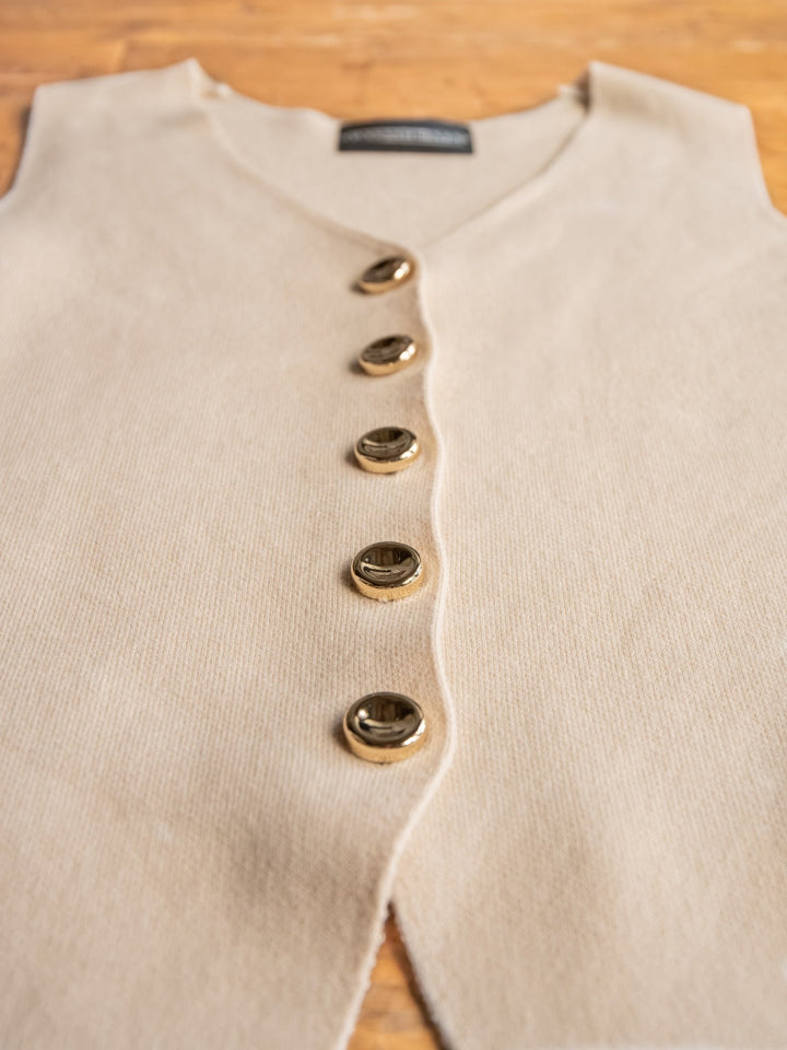 The Celara by Savannah Sevens western life{&}style is a beige sleeveless button-up top with elegant gold buttons, laid flat on wood. A black label appears at the neckline, highlighting the brand's sophisticated detail.