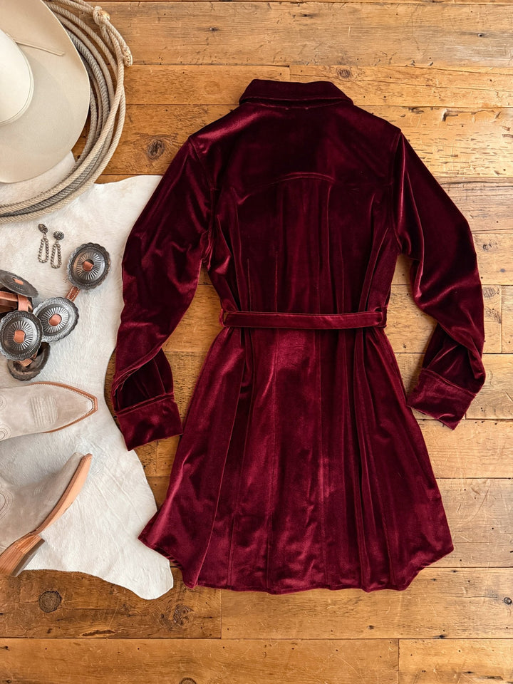 A Centralia Wine Velvet Button Up Dress by Savannah Sevens western life{&}style is laid flat on a wooden floor, surrounded by a white cowboy hat, silver jewelry, spurs, a white cowhide rug, and light-colored cowboy boots.