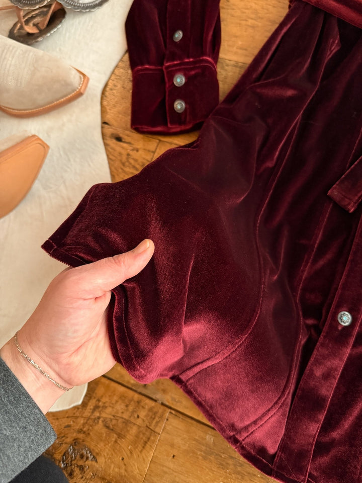 A hand holds the edge of the Centralia Wine Velvet Button Up Dress by Savannah Sevens western life{&}style, its deep burgundy velvet and metal buttons resting near cowboy boots and white fabric, evoking signature western style.