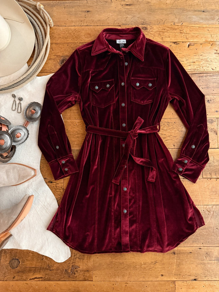 The Centralia Wine Velvet Button Up Dress by Savannah Sevens western life{&}style, with chest pockets and a waist tie, is styled on a wooden surface with a white hat, jewelry, and beige heeled boots on a white rug.