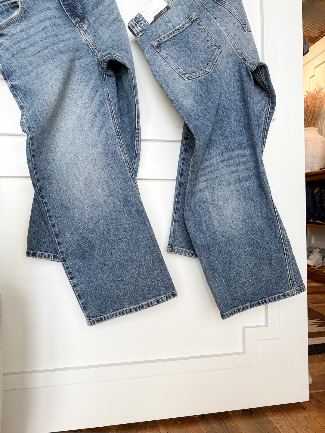 Two pairs of The Chainey Premier Rise Barrel Jean by Savannah Sevens western life{&}style hang side by side on a white paneled door, displaying medium wash denim with subtle fading on the thighs and seams. A wooden floor appears at the bottom.
