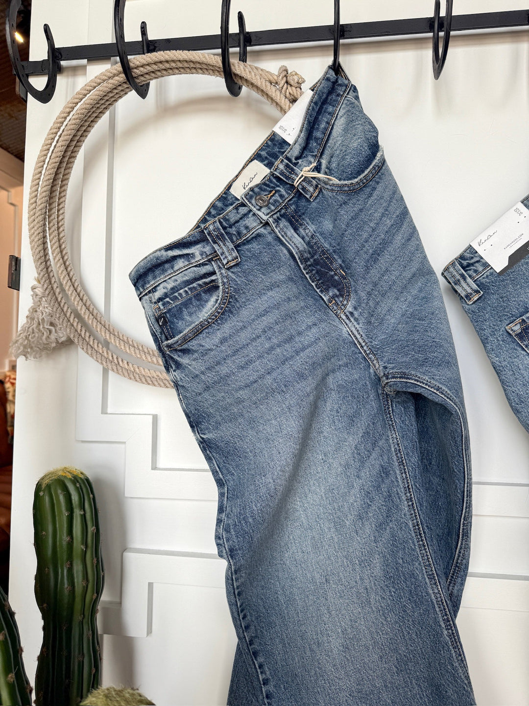 The Chainey Premier Rise Barrel Jean by Savannah Sevens western life{&}style hangs on a black hooked rack against a white wall, with a thick decorative rope and part of a cactus visible nearby.