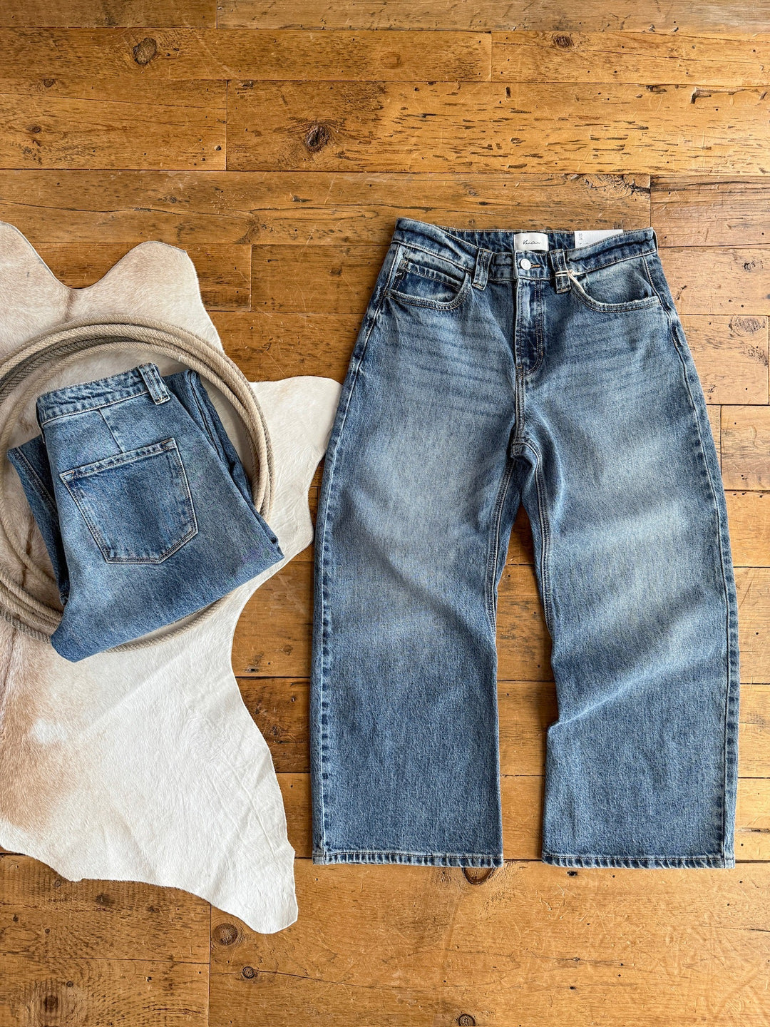 Two pairs of The Chainey Premier Rise Barrel Jean by Savannah Sevens western life{&}style on a wooden floor: one folded on a white cowhide rug with a rope, the other laid flat beside the rug, both in medium wash denim.