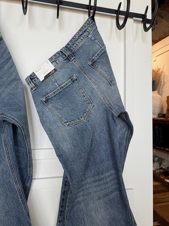 The Chainey Premier Rise Barrel Jean by Savannah Sevens western life{&}style, a medium wash denim, hangs on a black hook rack with a white tag on the waistband. In the background are a white door and shelves with decor items.