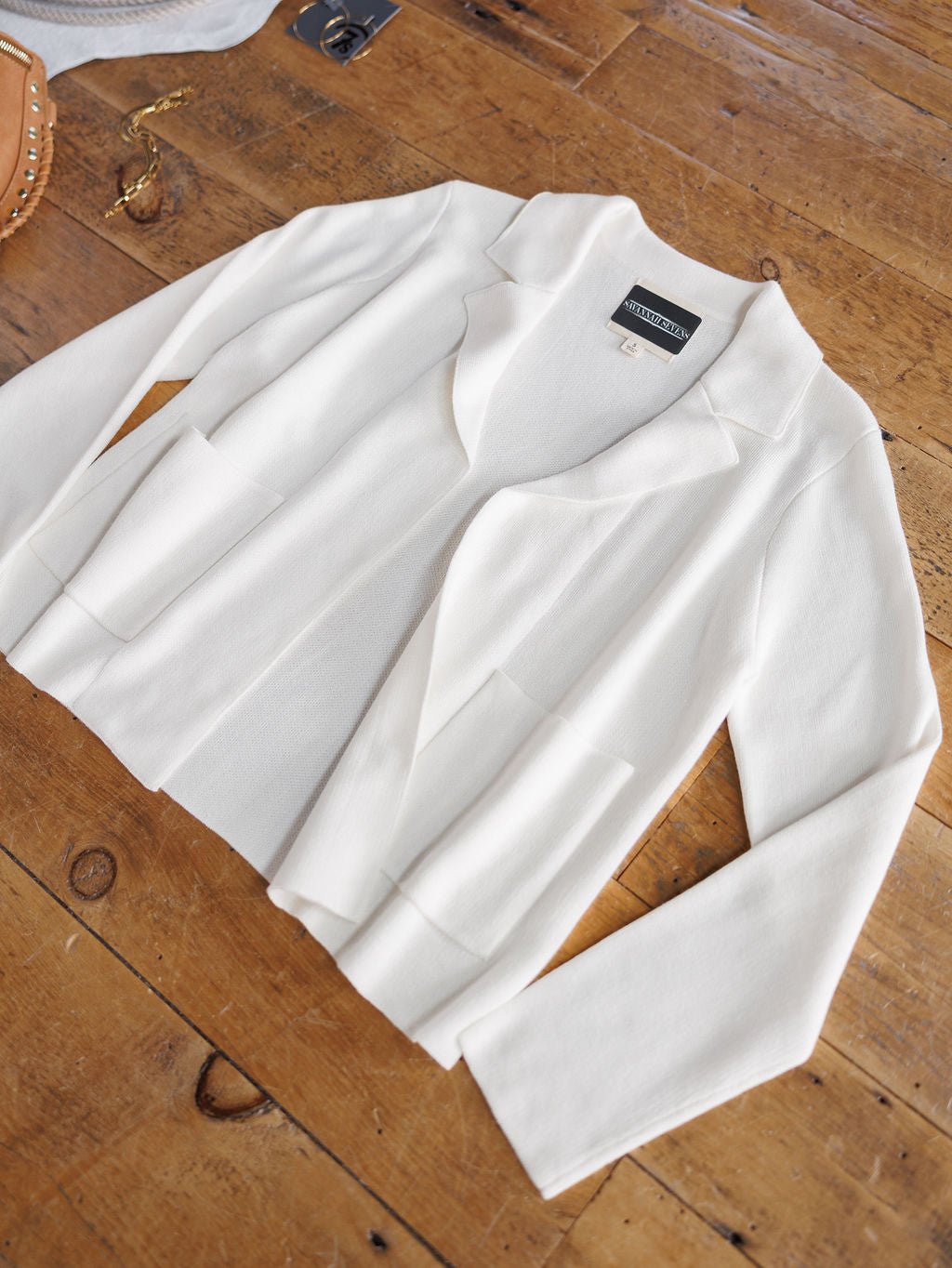 The Chamois White Cashmere Blend Cardigan by Savannah Sevens western life{&}style, a relaxed-fit, long-sleeve blazer-style piece with two front pockets and collar, lies flat on a wooden table beside a gold bracelet and brown handbag.