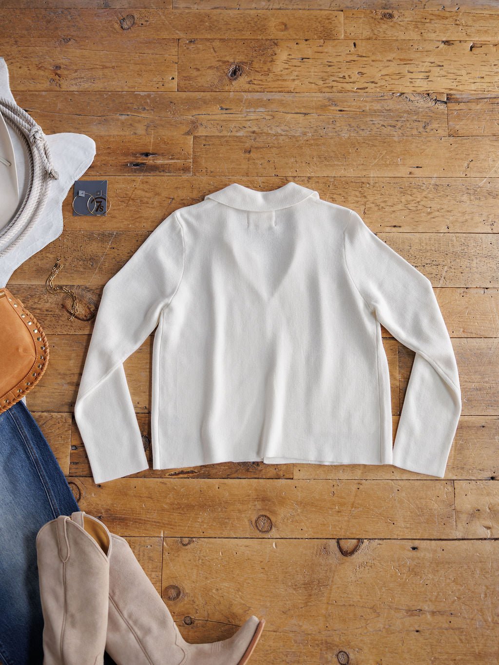 The Chamois White Cashmere Blend Cardigan by Savannah Sevens western life{&}style is laid flat on a wooden floor, paired with beige boots, denim jeans, a tan handbag, and a white hat.