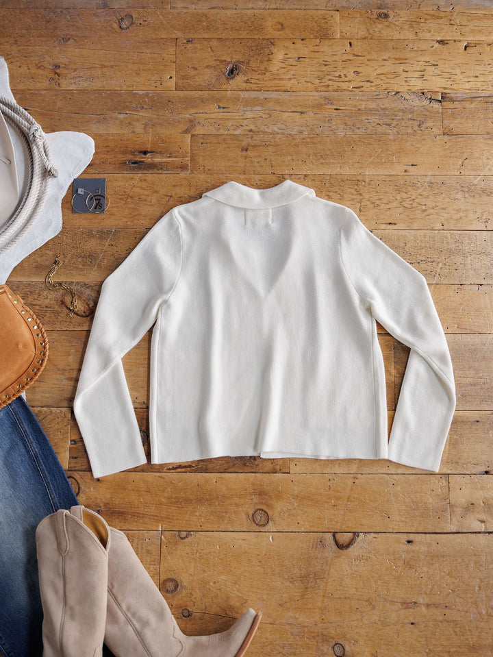The Chamois White Cashmere Blend Cardigan by Savannah Sevens western life{&}style is laid flat on a wooden floor, paired with beige boots, denim jeans, a tan handbag, and a white hat.
