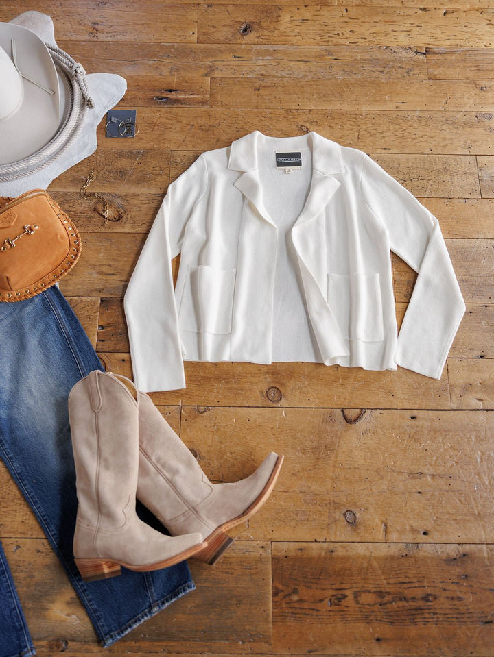 The Chamois White Cashmere Blend Cardigan by Savannah Sevens western life{&}style is displayed on a wooden floor with tan knee-high boots, blue jeans, a tan purse, and a white cowboy hat.