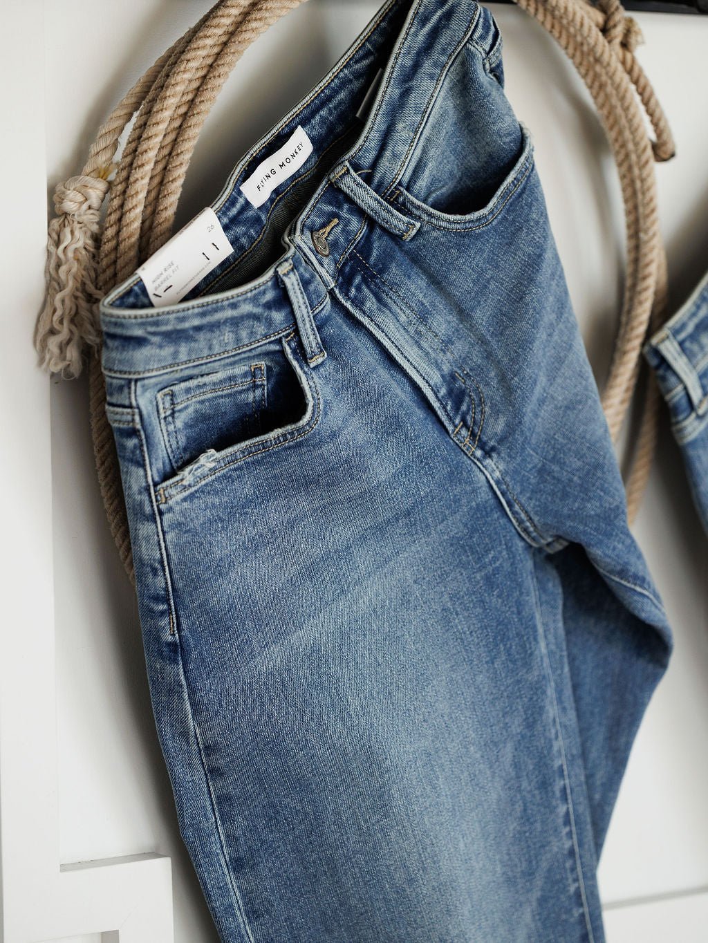 The Chase High Rise Barrel Jean by Savannah Sevens western life{&}style hangs on a hook with a coiled rope behind, featuring a medium wash, worn-in look, high rise fit, and a visible white label on the waistband.