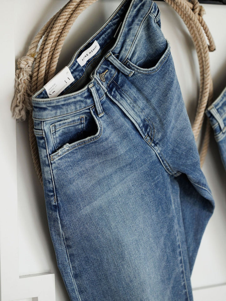 The Chase High Rise Barrel Jean by Savannah Sevens western life{&}style hangs on a hook with a coiled rope behind, featuring a medium wash, worn-in look, high rise fit, and a visible white label on the waistband.