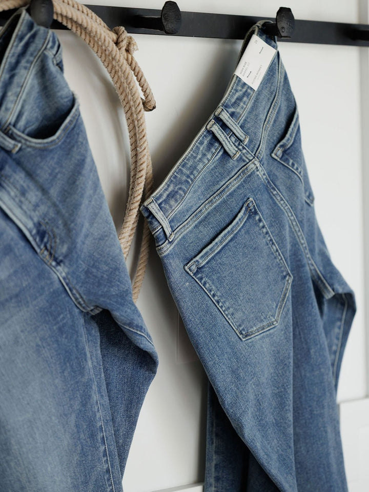 Two pairs of blue jeans, including The Chase High Rise Barrel Jean by Savannah Sevens western life{&}style and another medium wash style, hang on black hooks against a white wall with a coiled rope. One pair shows its back pocket and tag.
