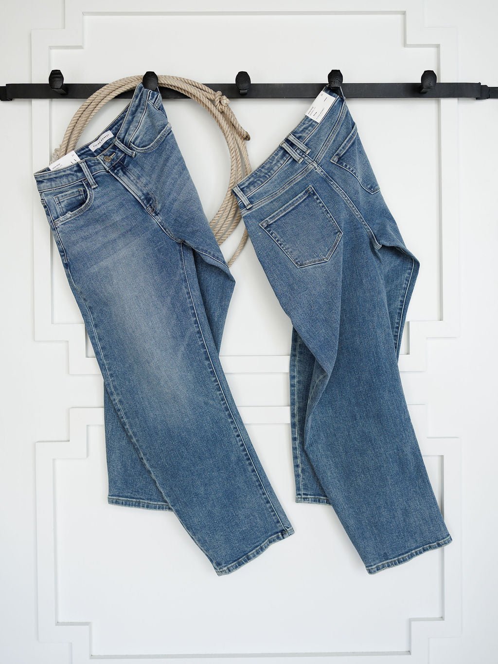 Two pairs of The Chase High Rise Barrel Jeans by Savannah Sevens western life{&}style hang side by side on a black rack against a white wall, with a thick rope looped behind them for a decorative touch.
