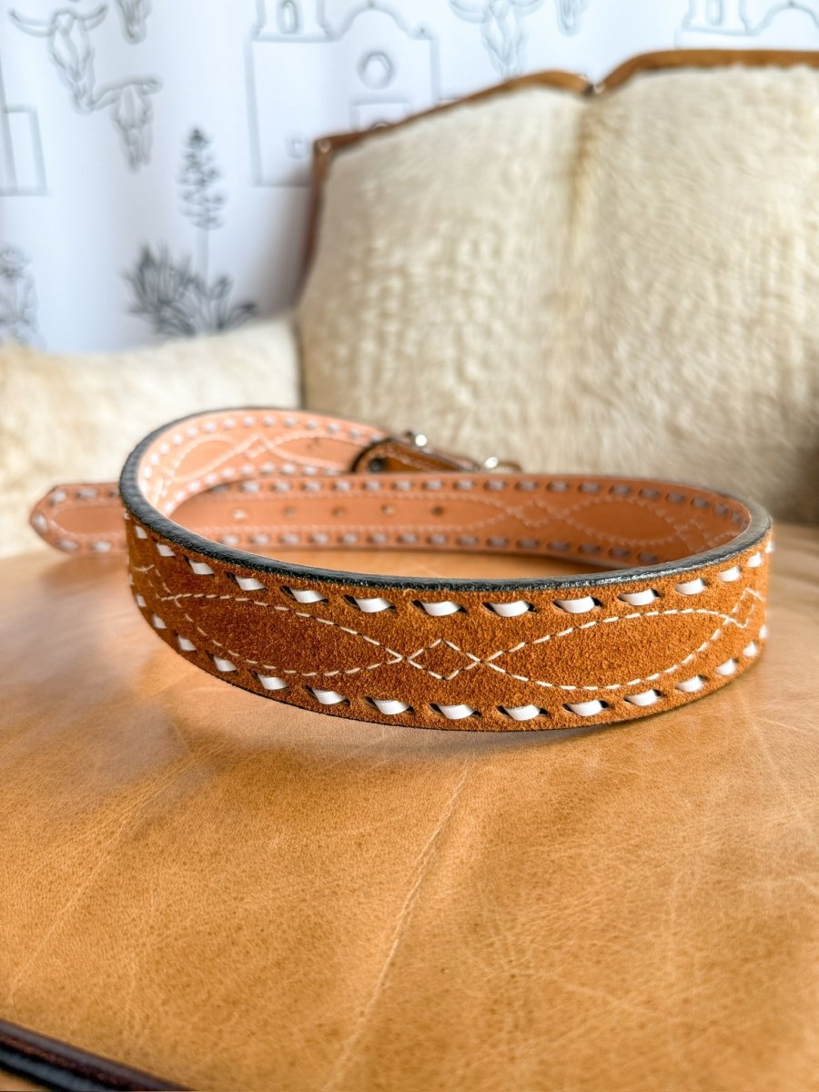 A close-up of the Chatham Belt by Savannah Sevens western life{&}style shows brown rough out leather with white stitching and decorative patterns, a sterling silver buckle, on tan leather beside a cream cushion and patterned wallpaper.