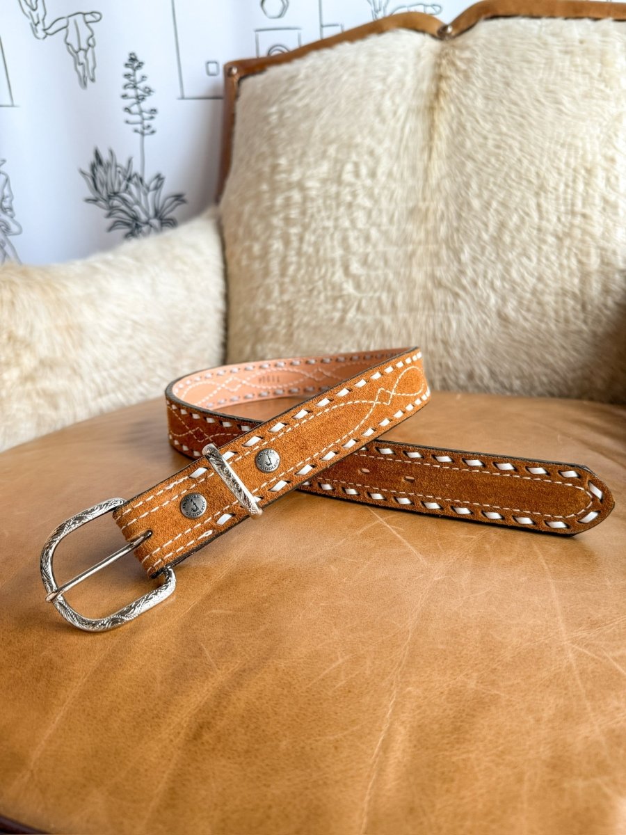 The Chatham Belt by Savannah Sevens western life{&}style, featuring white stitching and a sterling silver buckle, is coiled on a leather chair with a cream sheepskin cushion and a carved wooden backrest.