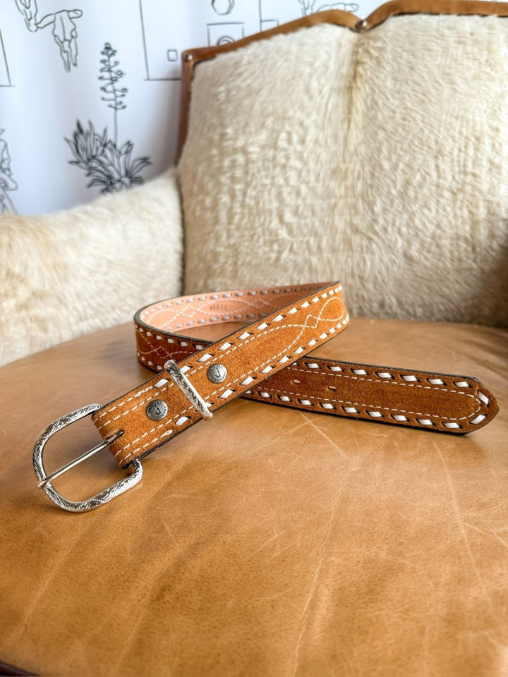 The Chatham Belt by Savannah Sevens western life{&}style, featuring white stitching and a sterling silver buckle, is coiled on a leather chair with a cream sheepskin cushion and a carved wooden backrest.