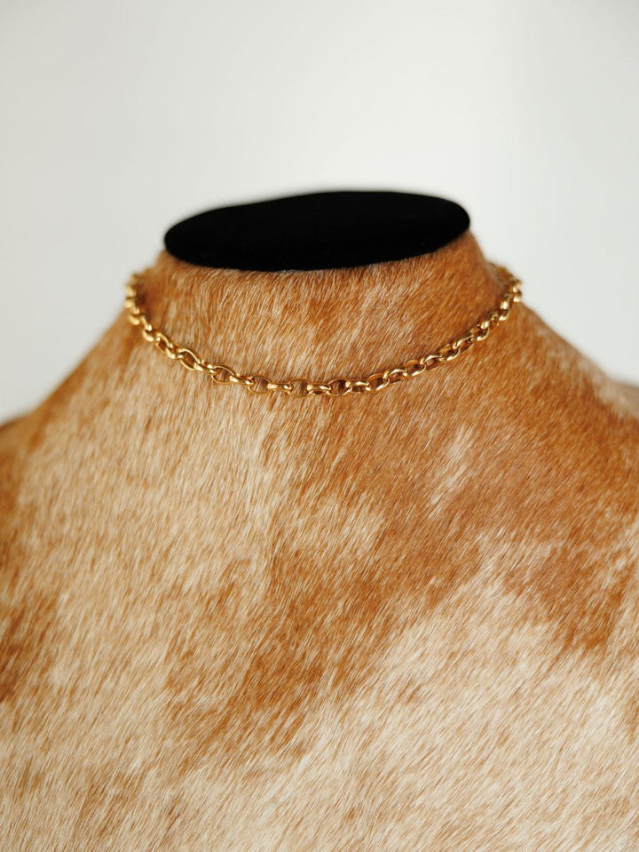 The Cheney Necklace by Savannah Sevens western life{&}style, a delicate gold choker, is displayed on a brown and white fur-textured bust with a black top against a plain white background.