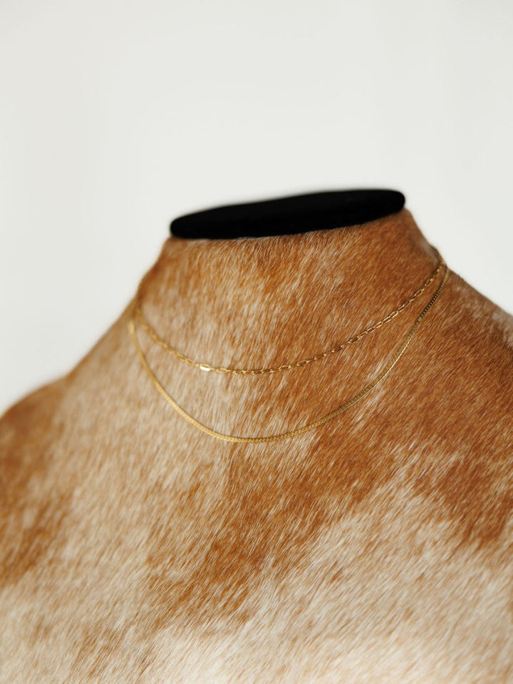 The Chenoa Necklace by Savannah Sevens western life{&}style features delicate, hypoallergenic gold chokers displayed on a brown-and-white speckled mannequin with a black top, set against a plain white background.