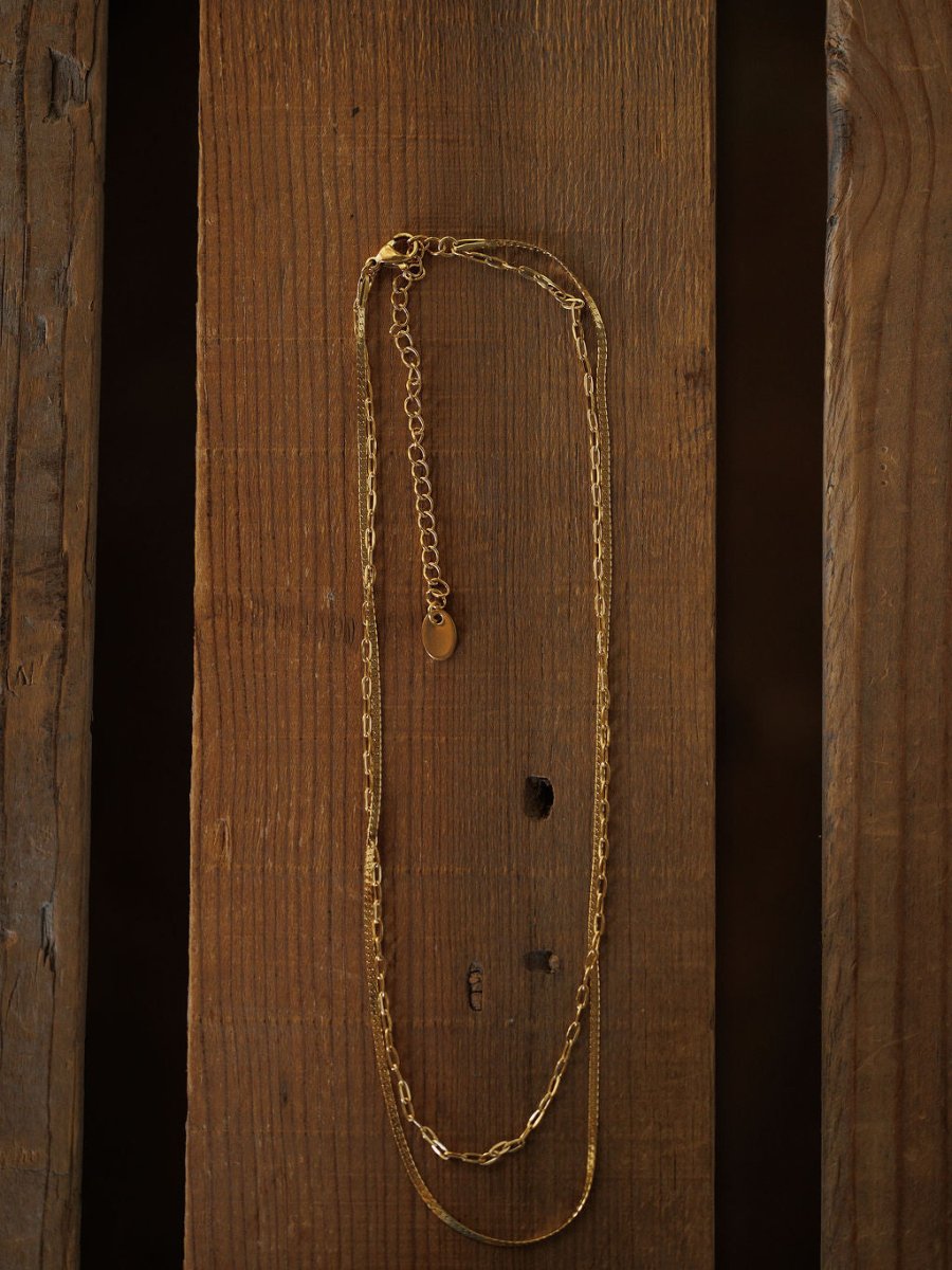 The Chenoa Necklace by Savannah Sevens western life{&}style features a delicate, hypoallergenic 18K gold filled chain and adjustable clasp, adding subtle elegance to any outfit. Displayed vertically against a rustic brown wooden background.