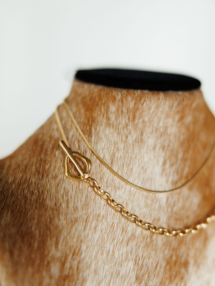 A close-up of two gold filled Cheraw Necklaces by Savannah Sevens western life{&}style, displayed on a tan furry mannequin bust. The dual chain design features thin and thick chains with a decorative toggle clasp.