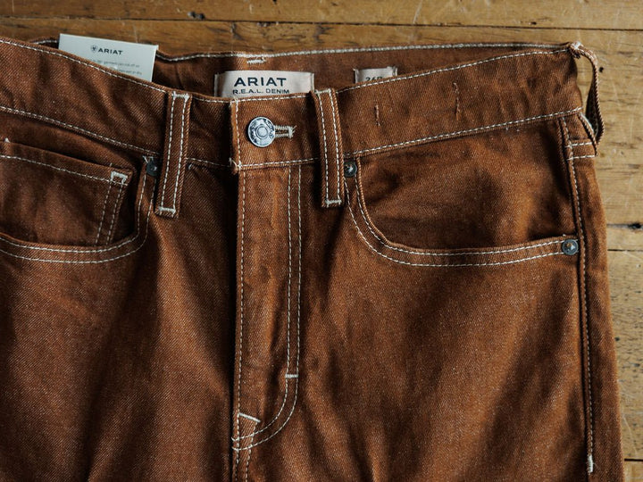 Close-up of The Chet High Rise Relaxed Straight Leg Jean by Ariat in brown sandstone denim with white stitching, silver button, and Ariat label on the waistband, displayed on a wooden surface.