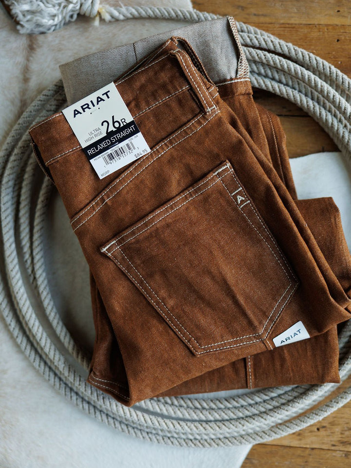 A pair of folded Ariat The Chet High Rise Relaxed Straight Leg Jeans, size 26, with a Relaxed Straight tag, are displayed on a wooden surface beside a coiled rope.