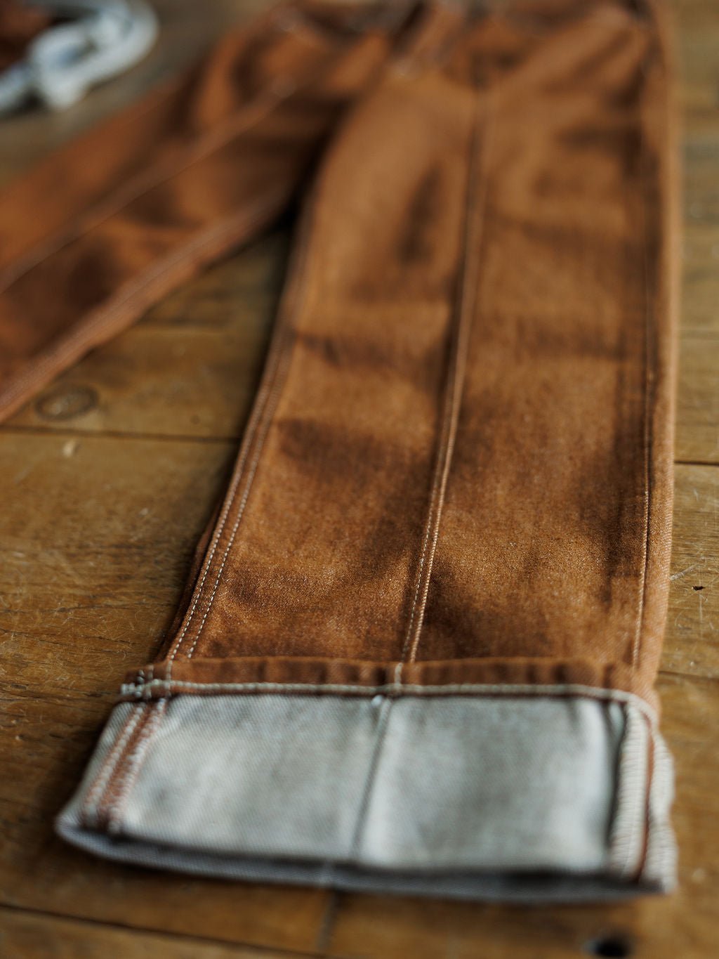 Close-up of The Chet High Rise Relaxed Straight Leg Jean by Ariat, featuring sandstone denim, visible stitching, and a folded cuff. Ultra high rise fit and seam details are clearly shown on the wooden surface.
