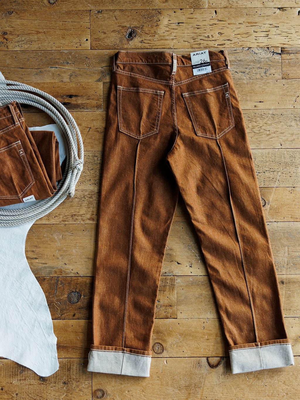 The Chet High Rise Relaxed Straight Leg Jean by Ariat in brown sandstone is laid flat on a wooden floor, with a coiled rope and folded jeans to the left, and a white cowhide partially visible in the lower left corner.