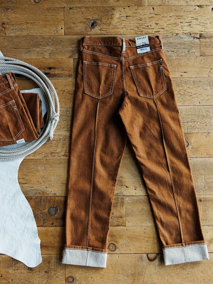 The Chet High Rise Relaxed Straight Leg Jean by Ariat in brown sandstone is laid flat on a wooden floor, with a coiled rope and folded jeans to the left, and a white cowhide partially visible in the lower left corner.