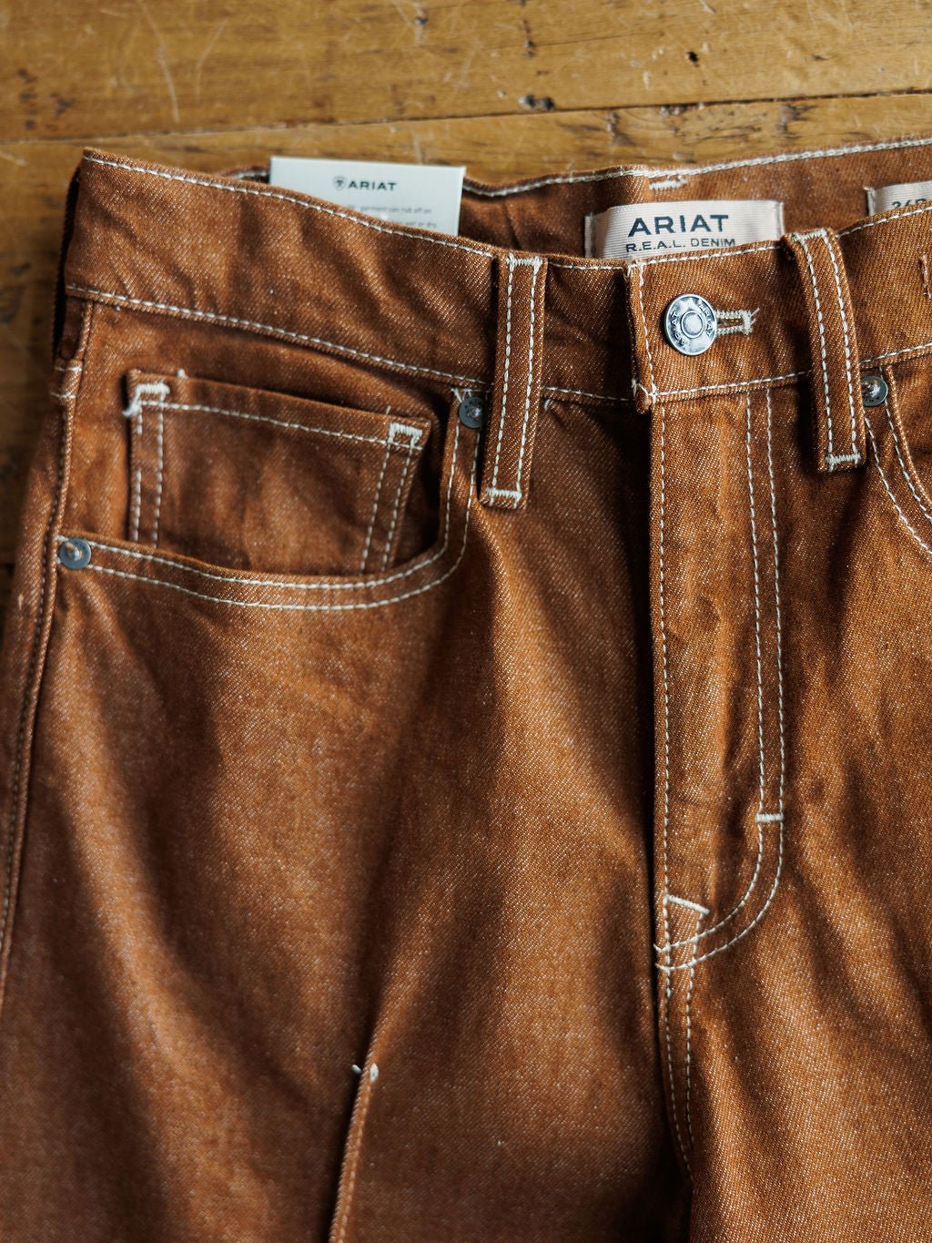 Close-up of Ariat's The Chet High Rise Relaxed Straight Leg Jean on wood, showing detailed stitching, pockets, belt loops, metal button, and Ariat label above the waistband. Ultra high rise with a relaxed straight leg fit.