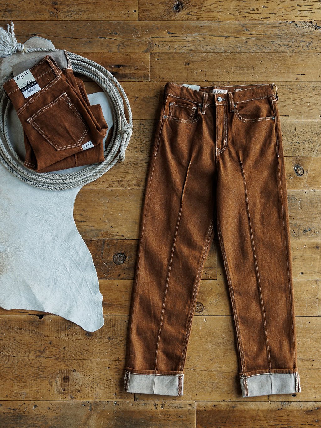 A pair of Ariat’s The Chet High Rise Relaxed Straight Leg Jeans in brown is laid flat on a wooden floor beside a white cloth and a round basket containing two folded sandstone denim jeans.