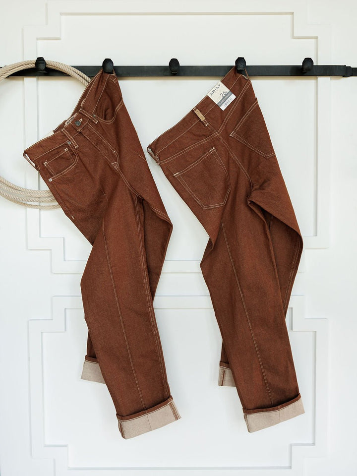 Two pairs of Ariat’s The Chet High Rise Relaxed Straight Leg Jeans in brown denim with rolled cuffs hang side by side on a black wall-mounted rod against a white wall with molding. A coiled rope hangs to the left.