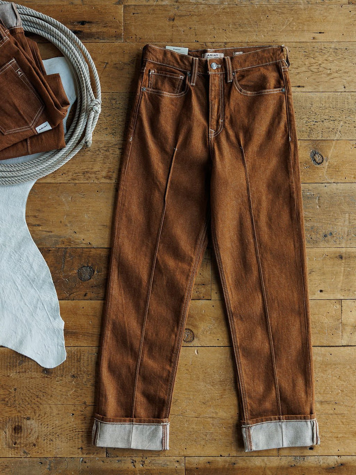 A pair of Ariat's The Chet High Rise Relaxed Straight Leg Jeans lies flat on a wooden floor, while another folded pair rests in a white basket partially covered by a white cloth.