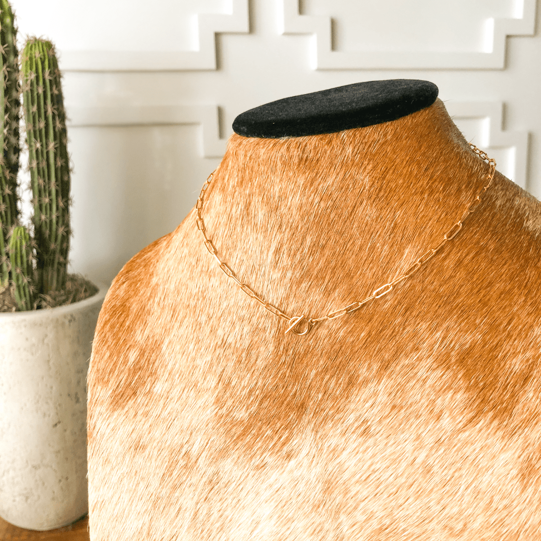 The Chetopa Necklace by Savannah Sevens western life{&}style is a 14K gold filled oval link necklace with a toggle clasp, shown on a cowhide bust, with a potted cactus and white decorative wall behind.