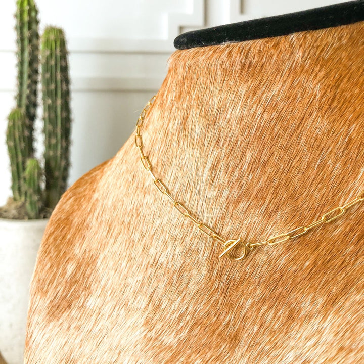 The Chetopa Necklace by Savannah Sevens western life{&}style, a 14K gold filled oval link design with toggle clasp, is shown on a tan and white cowhide jewelry bust with a potted cactus blurred in the background.
