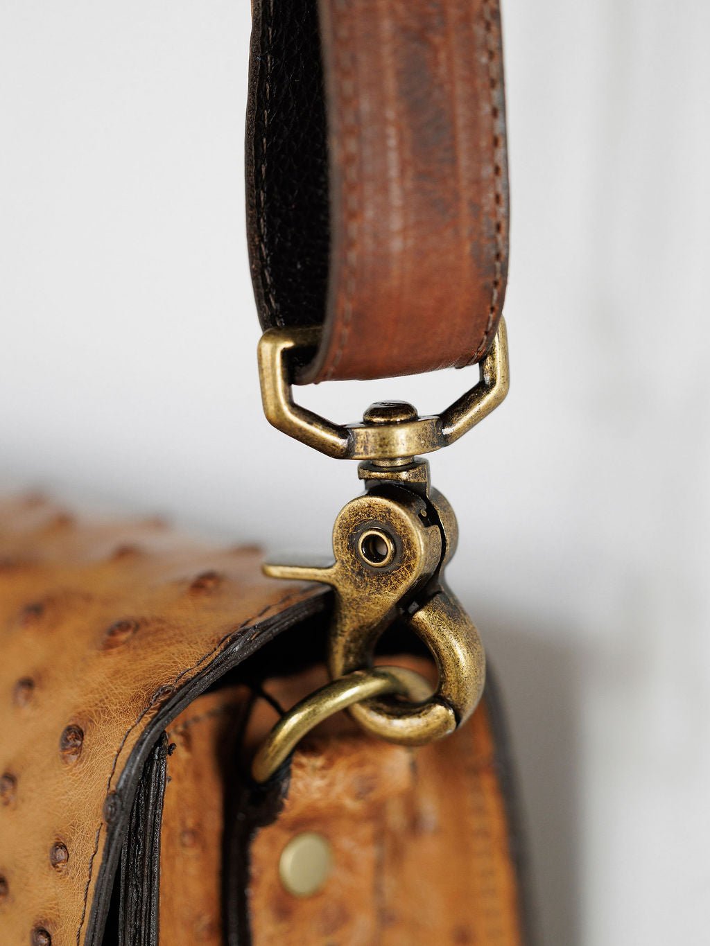 Close-up of a brass clasp securing the brown leather strap to the textured saddle tan Chicota Ostrich Leather Crossbody Clutch by Savannah Sevens western life{&}style, highlighting visible stitching and a rivet.