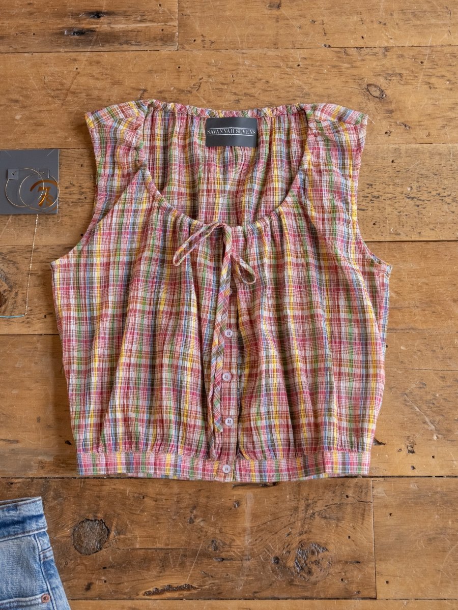 The Chikasaw Multi Colored Plaid Tank by Savannah Sevens western life{&}style is a sleeveless, cotton-blend crop top with pink, yellow, and green plaid and a neckline tie. Denim jeans and a gray item are partially shown beside it on a wooden surface.