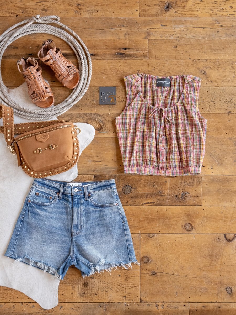 Flat lay of a summer outfit on wood: brown sandals, tan studded crossbody bag, Savannah Sevens western life{&}style Chikasaw Multi Colored Plaid Tank, light blue cropped denim shorts, rope, and hoop earrings in a box.