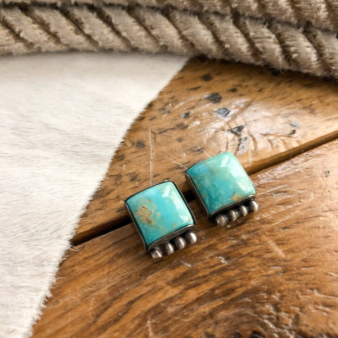 The Chino Valley Earrings by Savannah Sevens western life{&}style, featuring handmade turquoise and silver accents, rest on a wooden surface with soft fur and a coiled rope in the background.