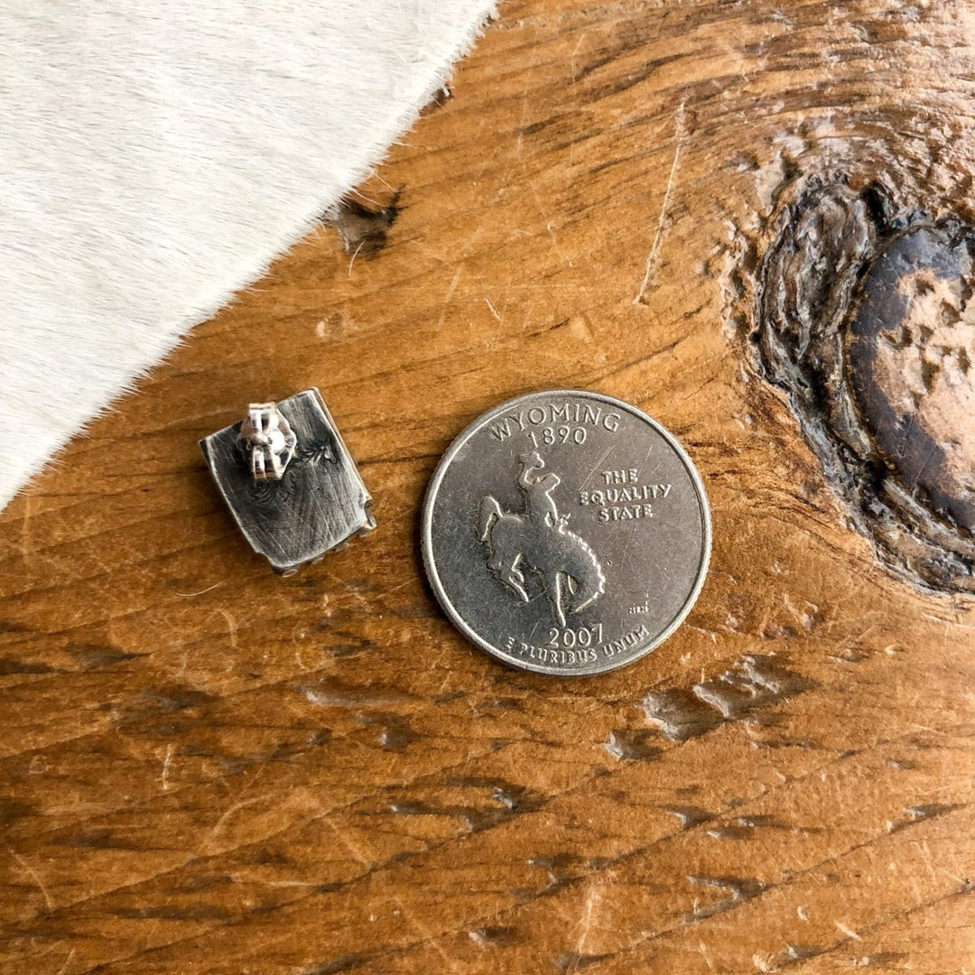 The Chino Valley Earrings by Savannah Sevens western life{&}style—a petite rectangular sterling silver stud —rests beside a 2007 Wyoming quarter on wood, with white fur accenting its western charm.