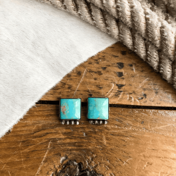 The Chino Valley Earrings by Savannah Sevens western life{&}style, handmade with Kingman turquoise and sterling silver accents, are displayed on wood beside a strip of rope and light cowhide.