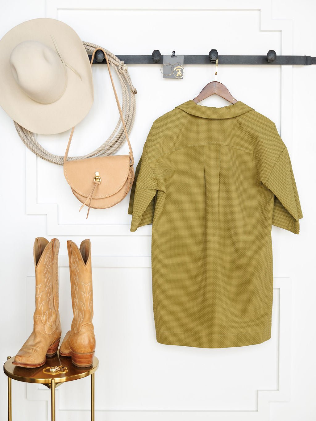 The Savannah Sevens western life{&}style Chino Valley Olive Textured Dress hangs on a wall hook beside a beige wide-brimmed hat and tan crossbody bag, with tan cowboy boots placed on a small round gold table below.
