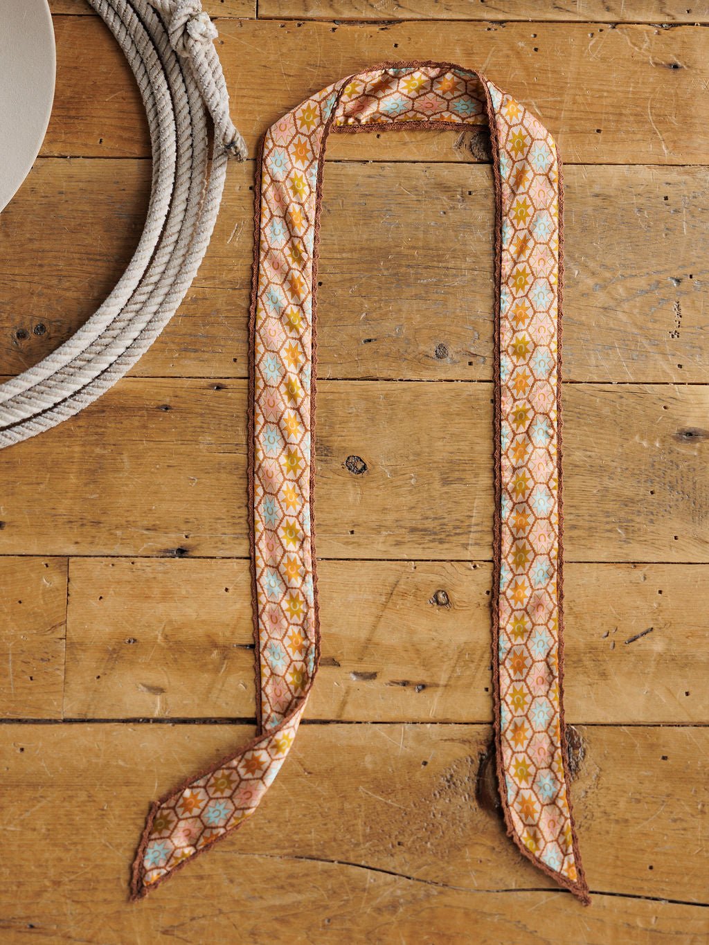 The Cimarron Twilly by Savannah Sevens western life{&}style, featuring orange, blue, and yellow diamond patterns on satin, is displayed on a rustic wooden surface beside a coiled white rope.