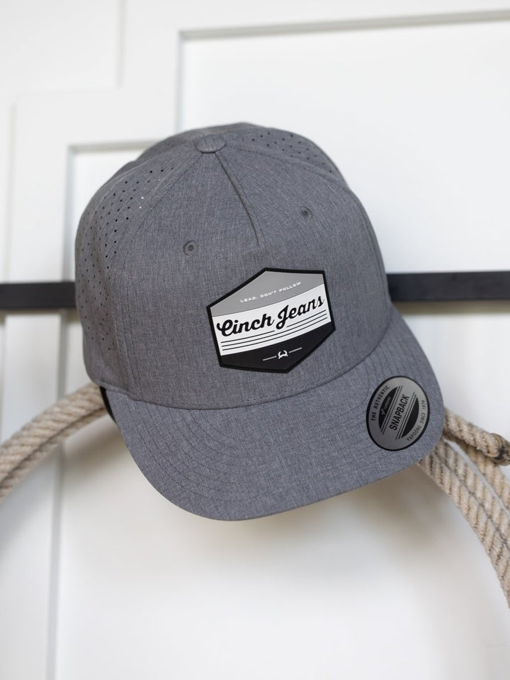A gray Cinch Lead Cap by Savannah Sevens western life{&}style, with a black and white logo, adjustable snapback, and thick rope around the hook, hangs on a white wall hook.